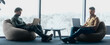 © Prostock-studio - Two men sit in bean bag chairs while working on laptops. They are focused on their screens. A large window shows a view of trees outside. Natural light fills the room.
