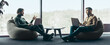 © Prostock-studio - Two men sit facing each other in an office space with large windows. They are working on laptops and having a discussion. The setting has a modern design with natural light coming in.