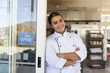 © wavebreak3 - Female chef standing at bakery door wearing white jacket, leaning with arms crossed, racks visible