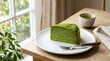 © Wanda - Delicate layers of vibrant green matcha crepe cake served on a white plate with a cup of tea beside it on a wooden table by a window