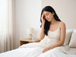 © Someone Cooked Here - indian young woman sitting on bed touching forehead representing headache migraine stress fatigue and home wellness concept