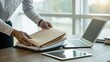 © nunoi - Person organizing paperwork at a desk with a laptop and tablet in a bright office setting.