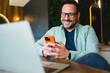 © djile - Smiling man using smartphone while sitting with laptop in cafe