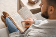 © Pixel-Shot - Young man reading book on grey sofa at home, closeup