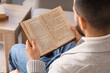 © Pixel-Shot - Young man reading book on grey sofa at home, closeup