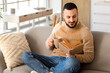 © Pixel-Shot - Handsome young man with cup of tea reading book on grey sofa at home