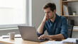 © Wiwat - Young man working on laptop at home desk with coffee and documents, focused and thoughtful expression in bright modern office
