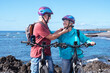 © luciano - Happy caucasian senior couple wearing helmets before riding on the beach with electric bicycles. Authentic elderly retired life concept. Horizon over sea