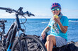 © luciano - Smiling active senior woman with bicycle sitting resting along a scenic seaside coastline eating an apple. Wearing a helmet and casual sportswear, enjoying a healthy outdoor lifestyle and the freedom