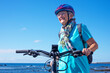 © luciano - Active senior woman cycling along a scenic seaside coastline on a sunny day. Wearing a helmet, eyeglasses, and casual sportswear, enjoying a healthy outdoor lifestyle and the freedom