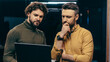 © Prostock-studio - Two men are engaged in a work discussion in an office. They are looking intently at a laptop screen. One man has curly hair and a green sweater, while the other has short hair and a yellow shirt.