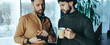 © Prostock-studio - Two men stand in an office environment. One man points at a phone while the other looks on with a cup in his hand. They appear to be engaged in conversation about something on the screen.