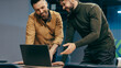 © Prostock-studio - Two men are looking at a laptop in an office setting. They are standing beside a table while talking and smiling. The environment has bright lights and a modern design.