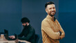 © Prostock-studio - A man stands confidently in an office setting with a warm smile. In the background, a coworker is focused on a laptop. The workspace has a modern design.