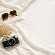 © zubayer - Minimalist Luxury Travel Flat Lay with Vintage Camera, Aviator Sunglasses, and Dried Palm Leaf on White Silk Fabric