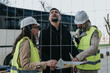 © qunica.com - Three construction professionals review a site blueprint while wearing hard hats and high-visibility vests. The engineers inspect plans at an outdoor construction site behind a safety fence.