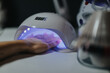 © qunica.com - A woman's hand with pink gel nails is placed under an LED nail lamp for curing. The scene shows a salon manicure in progress with purple-blue curing light.
