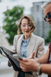 © qunica.com - A young businesswoman in glasses reviews a notebook while pointing and discussing with a colleague. They stand outdoors in an urban setting as she explains information from her folder.