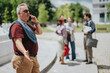 © qunica.com - A middle-aged business professional talks on his phone and gestures while standing outdoors. He speaks animatedly as a small group of colleagues converse in the blurred background.
