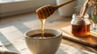 © nunoi - Honey drips from a wooden dipper into a ceramic cup filled with tea on a linen cloth, with a glass jar of honey and dried flowers blurred in the background.
