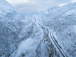 © AmazingAerialAgency - Aerial view of a winding road snaking through a vast snow-covered mountain valley with sparse winter trees under an overcast sky in Husoy, Troms, Norway.