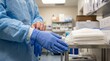 © Tatyana - Medical professional in blue gown and gloves prepares to wear protective gear in a clinical setting with surgical masks on a stainless steel table