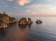 © AmazingAerialAgency - Aerial view of the Faraglioni sea stacks and a historic stone tower on a cliff at sunset in Scopello, Sicily, Italy.