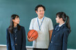 © buritora - A male teacher holding a basketball and a female student in a blazer are standing in front of the classroom blackboard.