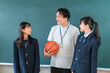 © buritora - A male teacher holding a basketball and a female student in a blazer are standing in front of the classroom blackboard.
