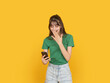 © Designerant - Smiling young woman looking at smartphone and covering mouth with hand, reacting to funny or surprising message concept. Isolated on bright yellow background with copy space for advertising use.