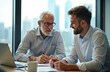 © Pete - Two men in formal shirts collaborate at a table with laptop and papers. Older man with glasses writes, younger man listens attentively. Office window backdrop shows city skyline.