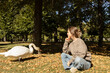 © Anna - cute adult girl in casual clothes sits on fallen leaves on a sunny day and happily looks at a large swan standing nearby. A nice walk in the park, an interesting meeting