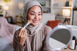 © Jovo Jovanovic/Stocksy - Woman in hijab applies makeup with brush, smiling.