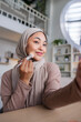 © Jovo Jovanovic/Stocksy - Woman in hijab applies makeup, looking in mirror.