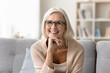 © fizkes - Portrait of happy mature woman wear eyeglasses and casual clothes posing for camera seated on comfortable sofa at home, smile, relaxing alone in modern living room with positive and joyful expression