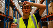 © Vali - Exhausted female warehouse worker wiping sweat while holding a water bottle. Employee wears a safety vest inside a distribution center. Heat stress concept for occupational health and safety.
