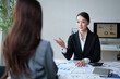 © MINAE - Businesswoman discussing financial strategy with a client in a modern office, explaining market analysis and reviewing corporate documents during a professional meeting