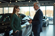 © miss irine - Mature salesman talks to woman customer in modern car dealership showroom. They discuss purchase options near black luxury automobile. Client considers new vehicle purchase.