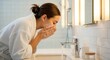 © Lucas - A woman in a white robe washing her face at a bathroom sink with soap and water