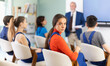 © JackF - Positive young woman in blue overalls sitting in class and looking back during teacher talk
