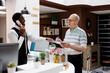 © DC Studio - Elderly retired caucasian guy signs reservation paperwork at counter of hotel lobby while african american employee takes a call. Senior customer fills out check-in papers as receptionist is on phone.