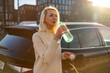 © vladim_ka - A woman drinks from a green plastic water bottle while standing next to a parked electric car charging. The sunlight creates a warm glow, highlighting her relaxed posture and casual attire in an urban