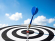 © ibrahim nuruz - A striking blue dart precisely hitting the bullseye of a dartboard symbolizing perfect aim strategic planning and ultimate achievement under an expansive blue sky with wispy clouds.