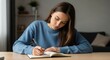 © Hasibnikon - Young woman writing in a notebook at a wooden desk in a cozy home office, focused on task