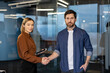 © Liubomir - Two business professionals woman with tablet and man shake hands and smile in a modern office, sealing a partnership and signaling collaboration, trust, and success