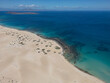 © AmazingAerialAgency - Aerial view of vast sand dunes meeting the turquoise Atlantic Ocean with scattered green shrubs under a clear blue sky in Lanzarote, Canarias, Spain.