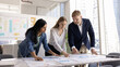 © fizkes - Three multiethnic teammates, marketing analysts gathered around conference desk in office, analyzing charts and graphs, reviewing campaign metrics, performance data in documents spread across table