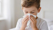 © pecherskiydotkz - Young boy blows his nose into a tissue in a close-up portrait, showing signs of mild illness or discomfort against a plain background.
