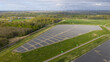 © AmazingAerialAgency - Aerial view of Zonnepark Braambergen solar farm with rows of photovoltaic panels surrounded by green fields, lush forests, and distant wind turbines Almere, Flevoland, Netherlands.