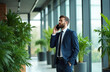 © Vadym - Bearded man in dark blue suit talks on cell phone. Stands in modern office with potted green plants near large windows. Engaged in business conversation, looking away from camera with hand in pocket,